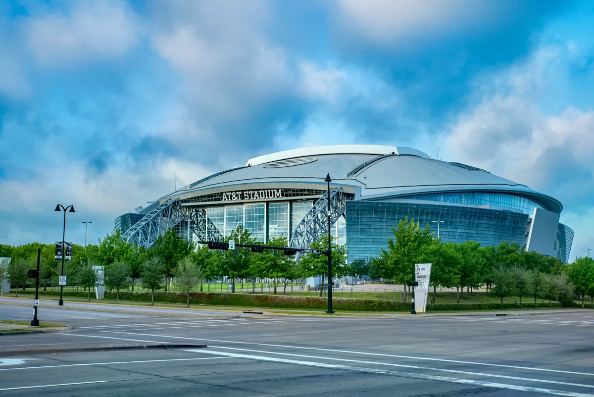 AT&T Stadium silver domed exterior with retractable roof and glass end walls surrounded by green trees in Arlington, Dallas