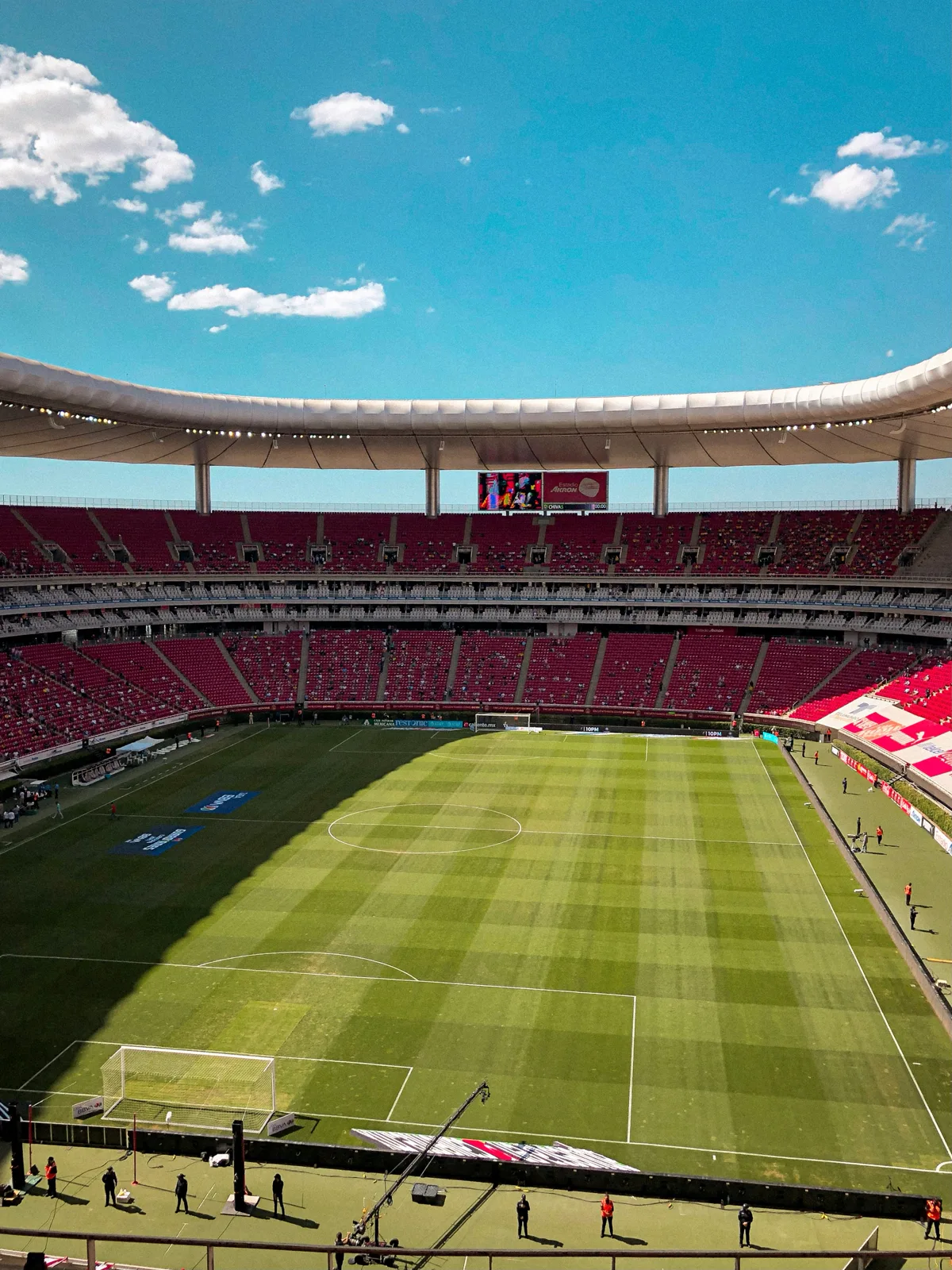 Estadio Akron interior before kick-off showing the pristine green pitch, vivid red seats, and open roof under a bright blue sky in Guadalajara
