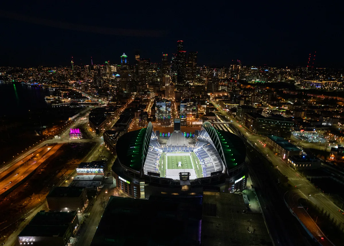 Lumen Field aerial night view with the illuminated green pitch at the centre and the glittering Seattle city skyline stretching behind it