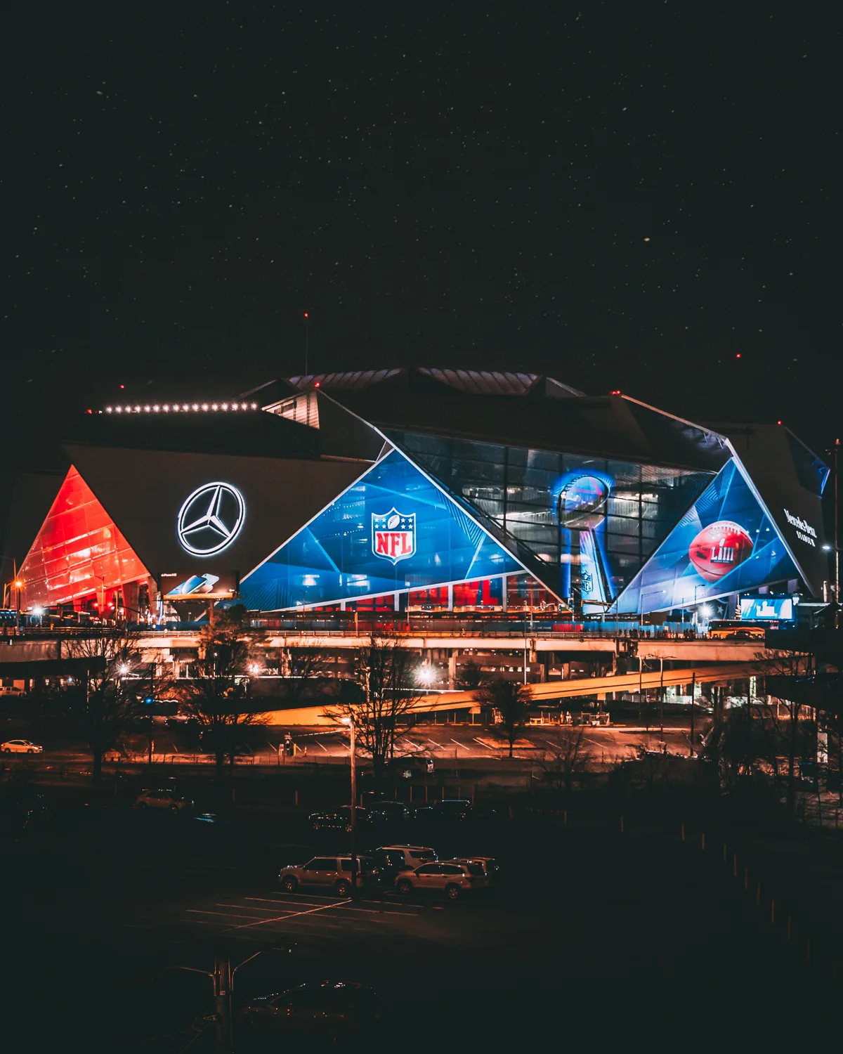 Mercedes-Benz Stadium exterior at night with its faceted diamond panels glowing blue and red under a star-filled Atlanta sky